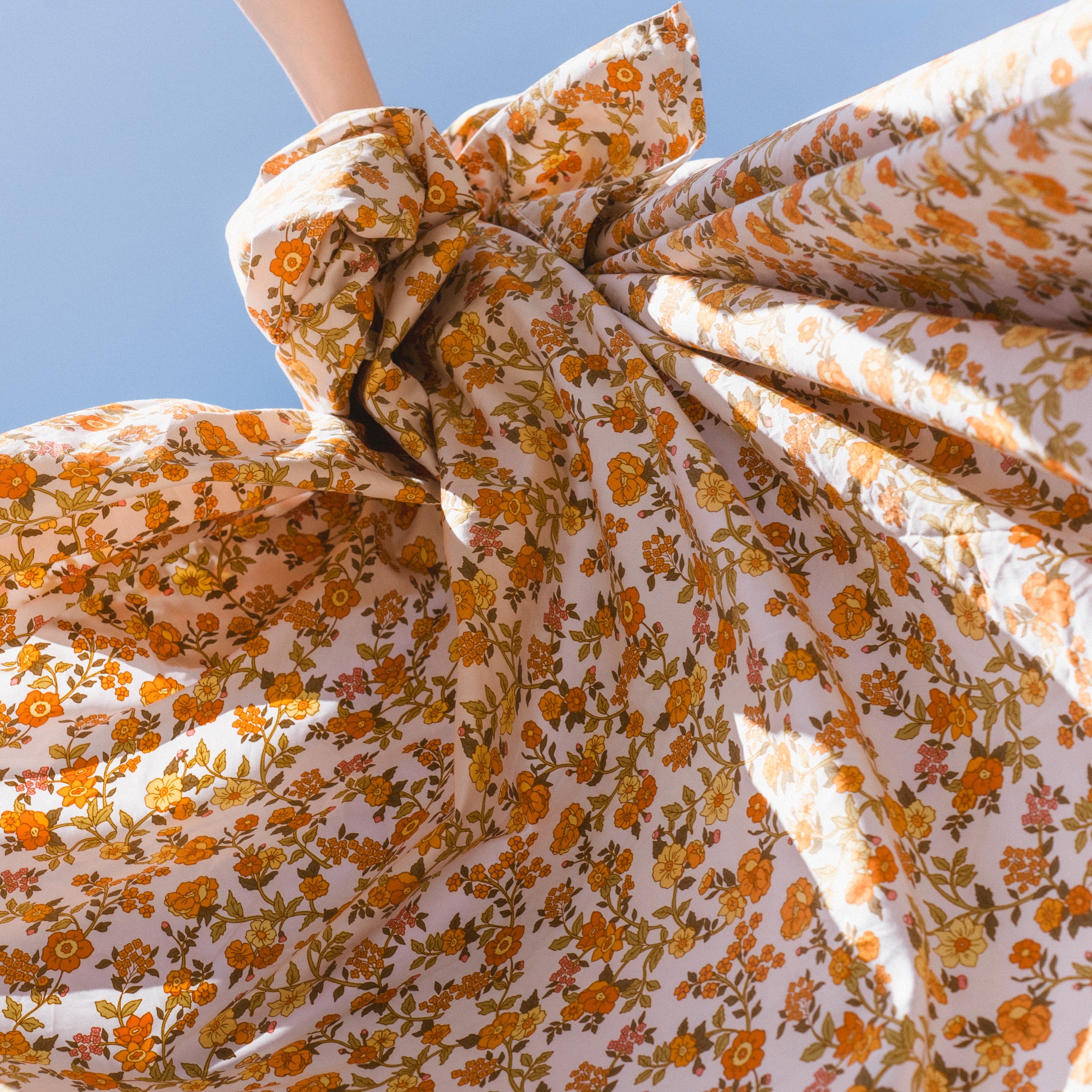 A beautiful cotton sheet being held up in the sunlight and showcasing it's pretty vintage floral that has pretty orange flowers with moss green, pops of pink and yellow. Beautiful sheets reminiscent of retro and vintage wallpaper from the sixties and seventies and fifties.